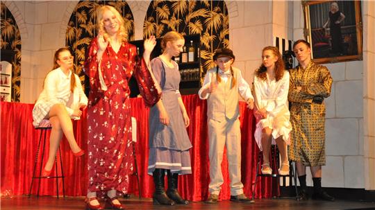 Sechs Personen in verschiedenen Kostümen auf einer Bühne vor rotem Vorhang und Wand mit Gemälden