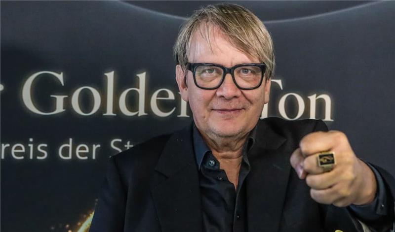 Freundeskreis Goldener Ton hofft auf Goslar-Konzert von Lea im Mai Ein Mann in dunklem Sakko und Hemd zeigt eine Hand mit einem auffälligen Ring vor einem Hintergrund mit der Aufschrift 'Goldener Ton'