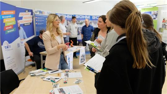 Frau mit braunem Pferdeschwanz hält Klemmbrett, steht vor Messestand mit mehreren Personen und Informationsmaterial auf Tisch.