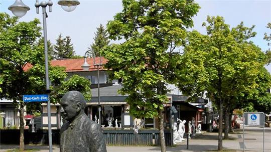 Bronzestatue eines Mannes in Anzug vor einem Platz mit Bäumen, Straßenlaternen und einem Gebäude mit rotem Dach