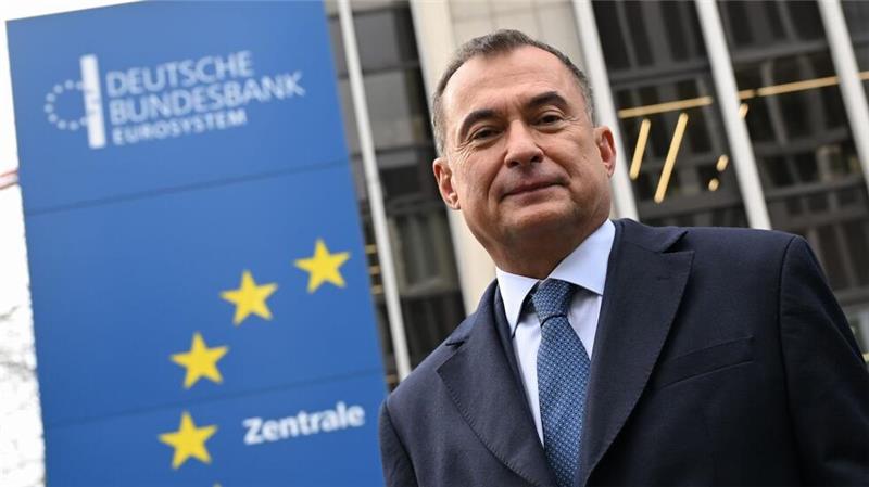 Mann im Anzug und Krawatte vor einem Gebäude mit einem blauen Schild der Deutschen Bundesbank und gelben Sternen