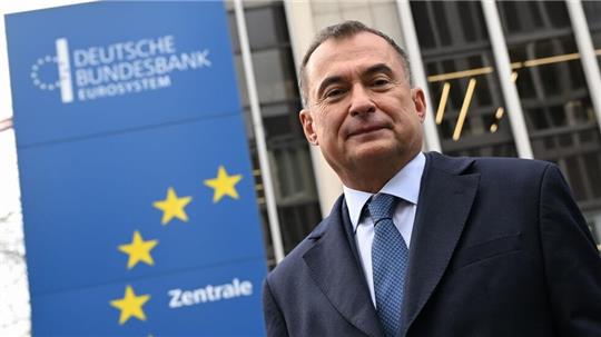 Mann im Anzug und Krawatte vor einem Gebäude mit einem blauen Schild der Deutschen Bundesbank und gelben Sternen