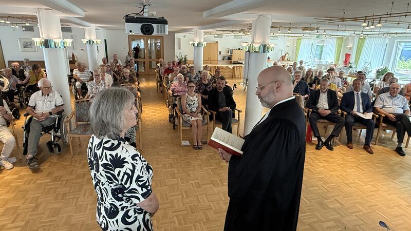 Ein Geistlicher  spricht mit einer Frau, im Hintergrund hören viele Menschen zu. 
