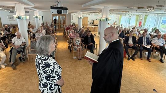 Ein Geistlicher  spricht mit einer Frau, im Hintergrund hören viele Menschen zu. 