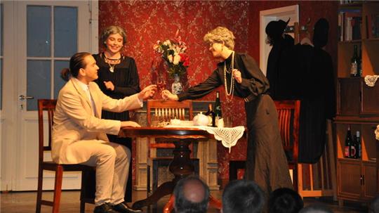 Theaterszene mit drei Personen in historischer Kleidung an einem Holztisch vor roter Tapete, eine Frau reicht einem sitzenden Mann einen Gegenstand