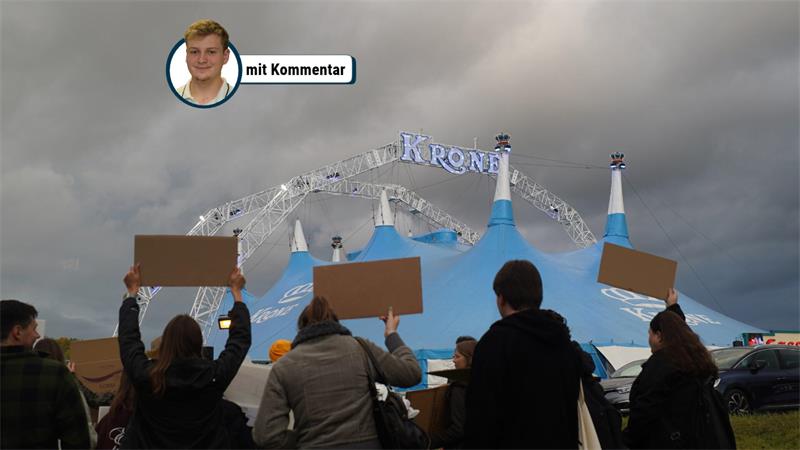 Das Bild zeigt eine Demonstration vor dem Zelt des Circus Krone. Mehrere Personen stehen im Vordergrund und halten Protestschilder in Richtung des Zirkuszeltes. Im Hintergrund ist deutlich das charakteristische blaue Krone-Zelt mit dem Schriftzug „KRONE“ zu sehen.