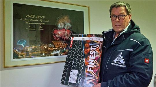Mann in dunkler Jacke hält zwei Feuerwerkskörperverpackungen vor einem gerahmten Bild mit Feuerwerk und Text.