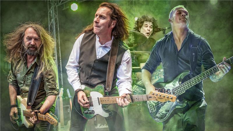 Vier Männer , drei mit Gitarre, einer im Hintergrund am Schlagzeug spielen ihre Instrumente im grünen Licht. 