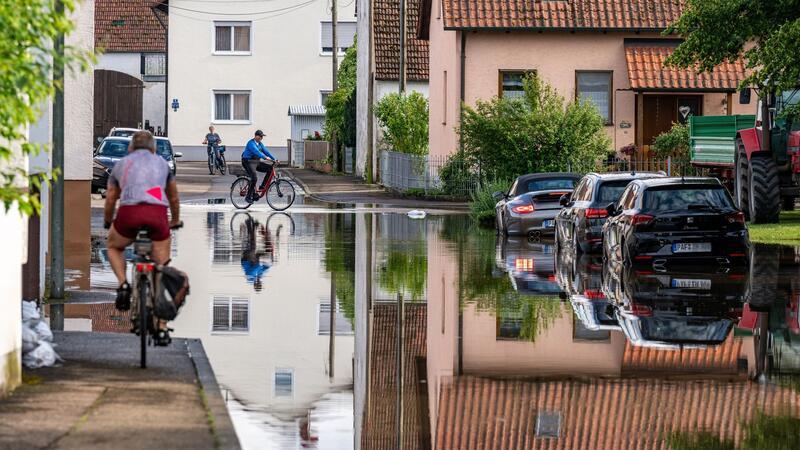 Radler fahren durch eine überflutete Straße. Im Bereich der Gemeinde Baar-Ebenhausen war ein Damm an zwei Stellen geborsten.