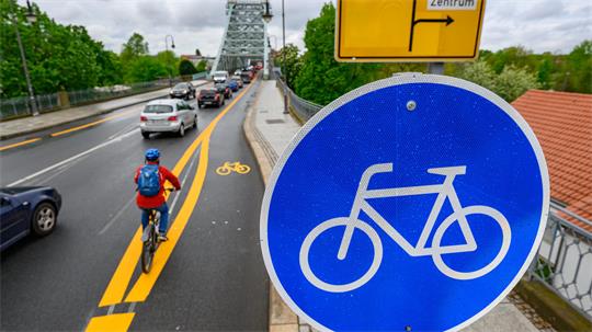 Fahrradfahrer auf einem markierten Radweg neben einer Straße mit Autos auf einer Brücke und einem blauen Verkehrsschild mit Fahrradsymbol