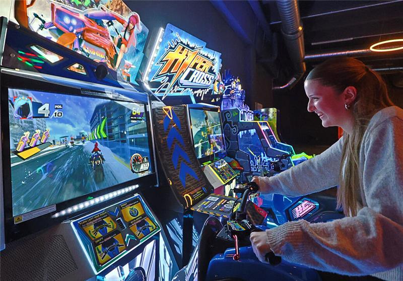 Junge Frau spielt an einem Motorrad-Rennspiel-Automaten in einer Spielhalle mit mehreren leuchtenden Spielautomaten.