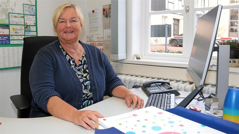 Ältere Frau mit blauem Cardigan sitzt an einem Schreibtisch vor einem Computer in einem hellen Büro