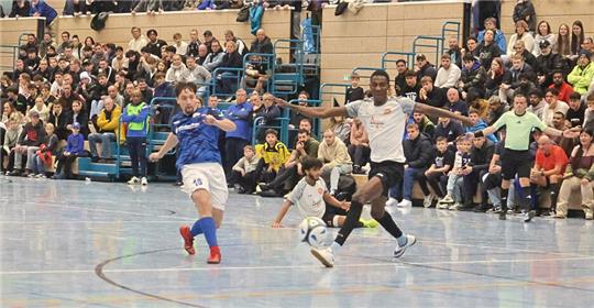 Drei junge Fußballspieler in Sportkleidung kämpfen um den Ball auf einem Hallenboden, im Hintergrund sitzen zahlreiche Zuschauer auf Tribünen