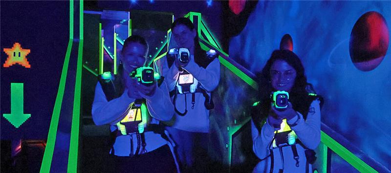 Drei Personen mit Laserpistolen und Westen in einer dunklen, neonbeleuchteten Lasertag-Arena