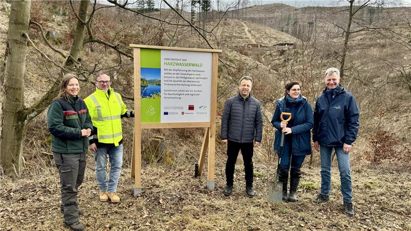 Fünf Personen stehen vor einem Informationsschild in einem kahlen Waldgebiet, eine Person hält eine Schaufel.