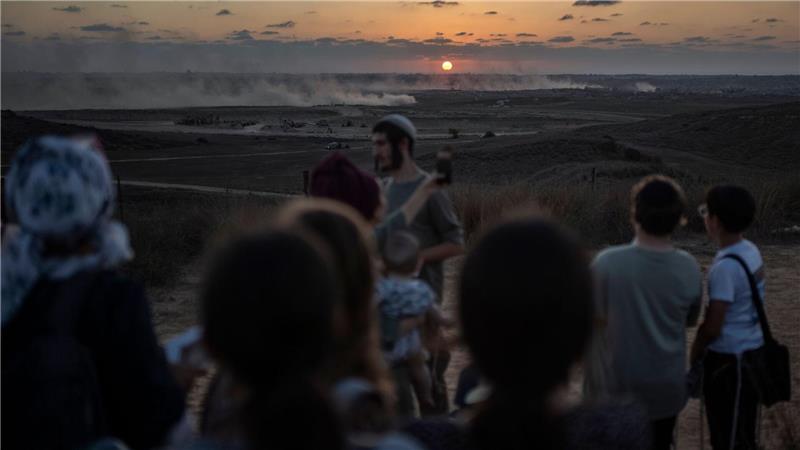 Bilder des Tages Rechtsgerichtete israelische Siedler versammeln sich an der Grenze zum Gazastreifen, während im Hintergrund Rauchschwaden von vorrückenden israelischen Panzern zu sehen sind.