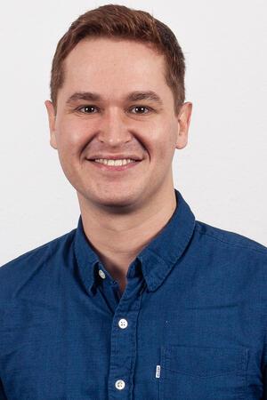 Ein Mann im blauen Hemd steht vor einer weißen Wand.