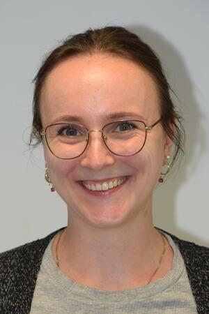 Person mit zusammengebundenem Haar, trägt Ohrringe und eine Halskette, bekleidet mit grauem Oberteil und dunklem Strickjacke