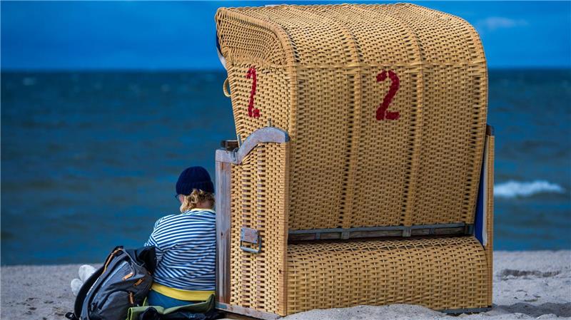 Regenschauer und kräftiger Wind an der Ostseeküste: Eine Frau sitzt mit Mütze bei wechselhaftem Wetter neben einem Strandkorb auf der Insel Poel.