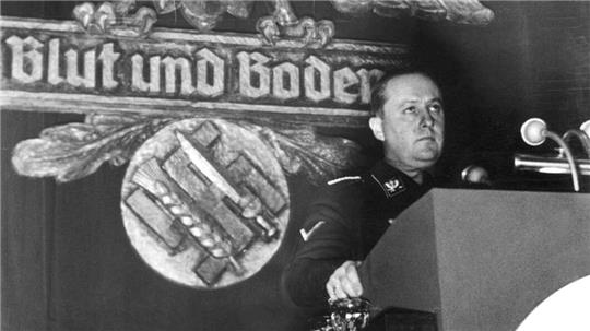 Mann in Uniform steht an Rednerpult vor Wand mit großem Adler und Schriftzug 'Blut und Boden'.