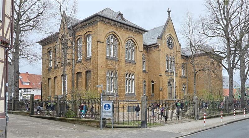 Zweistöckiges historisches Gebäude mit vielen hohen Fenstern an einer Straßenecke, umgeben von einem Metallzaun und kahlen Bäumen