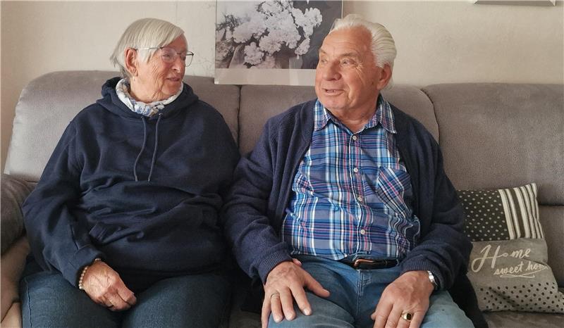 Zwei ältere Personen sitzen nebeneinander auf einem Sofa, im Hintergrund ein Bild mit Blumen und ein Kissen mit der Aufschrift 'Home sweet home'