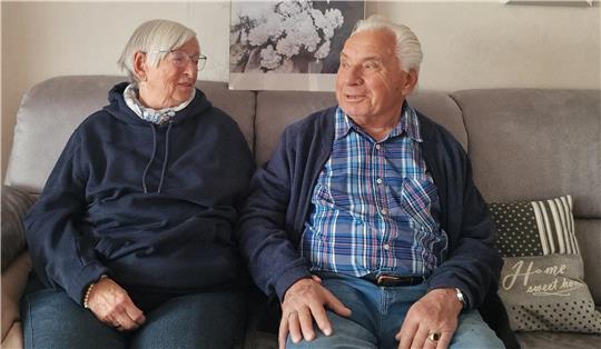 Zwei ältere Personen sitzen nebeneinander auf einem Sofa, im Hintergrund ein Bild mit Blumen und ein Kissen mit der Aufschrift 'Home sweet home'