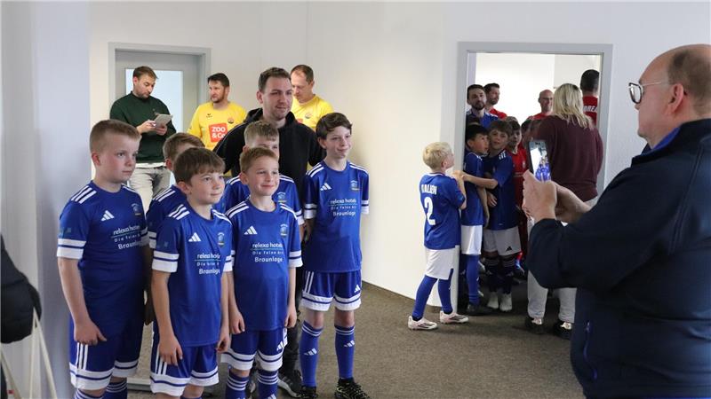 Gruppe von Kindern in blauen Fußballtrikots steht in einem Flur, einige Erwachsene im Hintergrund und ein Erwachsener im Vordergrund mit einem Smartphone in der Hand