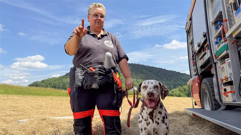 Person in Einsatzkleidung steht auf einem Feld neben einem Dalmatiner vor einem offenen Fahrzeug mit Ausrüstung