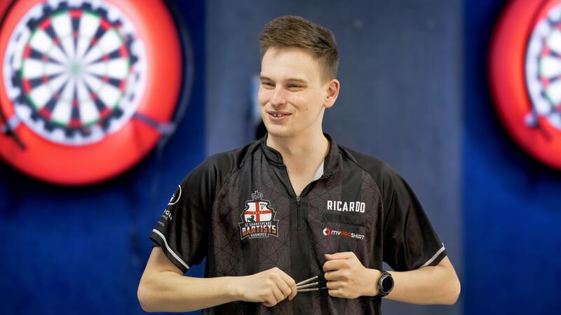 Junger Mann in schwarzem Dart-Trikot hält drei Darts in der Hand vor zwei Dartscheiben an der Wand