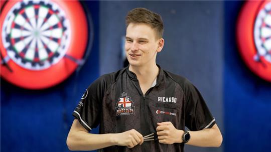 Junger Mann in schwarzem Dart-Trikot hält drei Darts in der Hand vor zwei Dartscheiben an der Wand