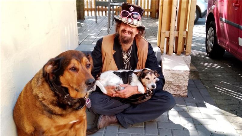 Mann sitzt auf dem Boden vor einer Mauer und hält einen kleinen Hund im Schoß, ein großer Hund sitzt daneben auf dem Boden.