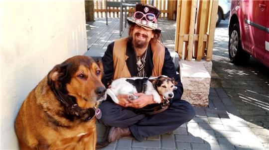 Mann sitzt auf dem Boden vor einer Mauer und hält einen kleinen Hund im Schoß, ein großer Hund sitzt daneben auf dem Boden.