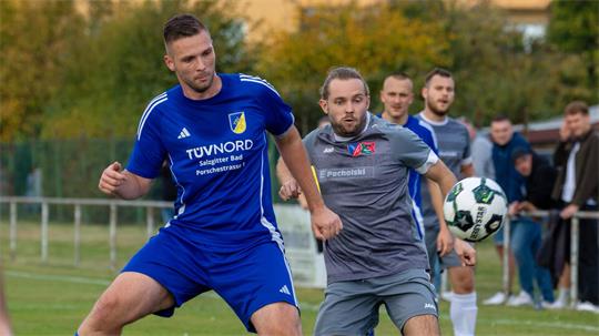 Zwei Fußballspieler in Aktion auf dem Spielfeld, einer in blauem Trikot mit Aufschrift 'TÜVNORD', der andere in grauem Trikot mit Aufschrift 'E Pocholski', im Hintergrund weitere Spieler und Zuschauer