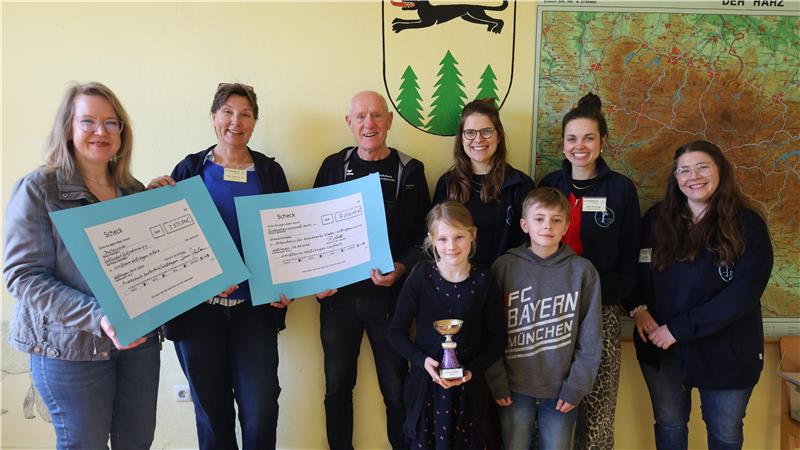 Gruppe von acht Personen steht vor einer Wand mit einer Landkarte und einem Wappen, zwei Personen halten große blaue Karten, ein Kind hält einen Pokal