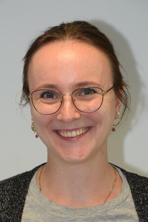 Frau mit braunem, zurückgebundenem Haar trägt grauen Pullover und goldene Ohrringe sowie eine dünne goldene Halskette vor hellem Hintergrund