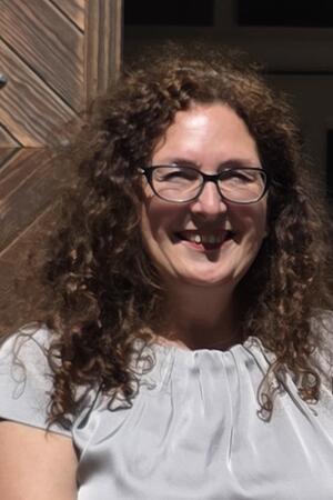 Person mit lockigem Haar und weißer Bluse vor Holzwand.