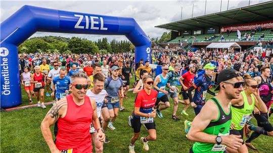 Läufer starten bei einem Laufwettbewerb unter einem blauen Zielbogen auf einem Sportplatz mit Zuschauern auf der Tribüne