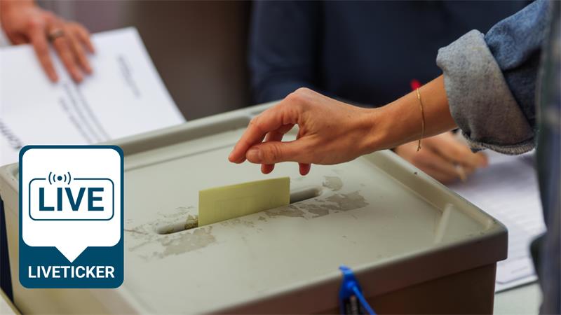 Hand, die einen Wahlzettel in eine graue Wahlurne steckt, im Hintergrund unscharfe Personen und Papiere