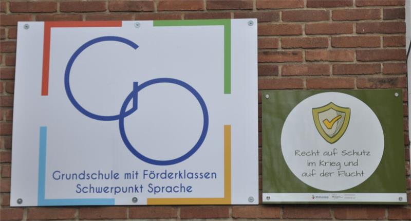Oker drückt aufs Tempo: Millionenschwere Module rücken an Zwei nebeneinander an einer Backsteinwand befestigte Schilder, links mit stilisiertem Logo und Text "Grundschule mit Förderklassen Schwerpunkt Sprache", rechts mit Schildsymbol und Text "Recht auf Schutz im Krieg und auf der Flucht"