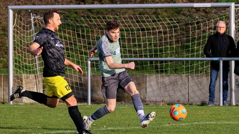 Zwei Fußballspieler in Aktion auf dem Spielfeld, einer in schwarzem Trikot mit gelber Hose, der andere in grauem Trikot, ein bunter Ball vor ihnen.