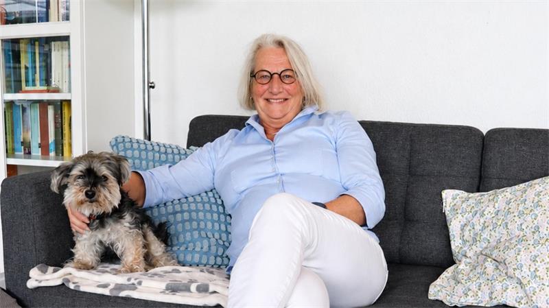 Ältere Frau mit Brille und hellblauer Bluse sitzt auf grauem Sofa neben kleinem Hund und lehnt sich an Kissen.