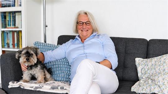 Ältere Frau mit Brille und hellblauer Bluse sitzt auf grauem Sofa neben kleinem Hund und lehnt sich an Kissen.