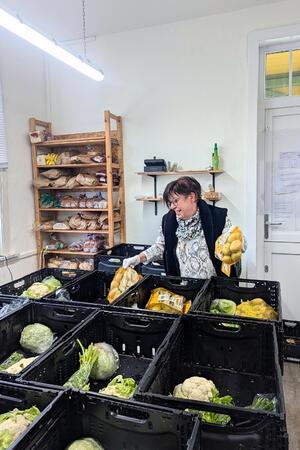 Sabine Krause befült Kisten und steht zwischen Gemüse, Brot und weiteren Lebensmitteln.