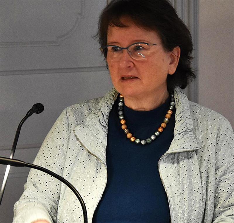 Frau mit dunklem Oberteil und hellgrauer, strukturierter Jacke sowie bunter Perlenkette vor grauer Wand und Mikrofon