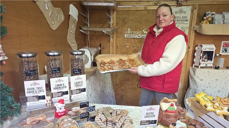 Verkaufsstand mit verschiedenen Hundekeksen in Tierform, darunter Knochen und Pfoten, sowie drei Behältern mit Trockenfutter und weihnachtlicher Dekoration.