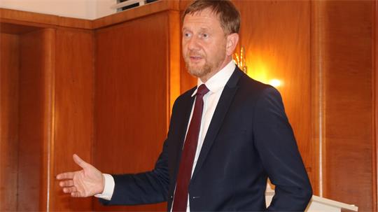 Mann mit Bart und Anzug gestikuliert mit linker Hand in einem holzvertäfelten Raum mit warmem Licht