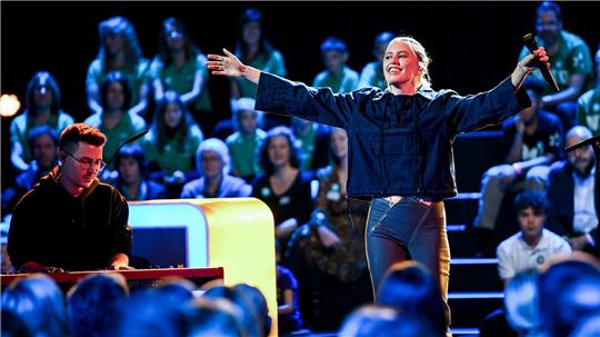 Person mit ausgestreckten Armen hält Mikrofon auf einer Bühne vor Publikum, eine weitere Person spielt Keyboard im Vordergrund