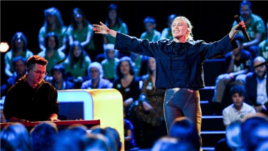 Person mit ausgestreckten Armen hält Mikrofon auf einer Bühne vor Publikum, eine weitere Person spielt Keyboard im Vordergrund