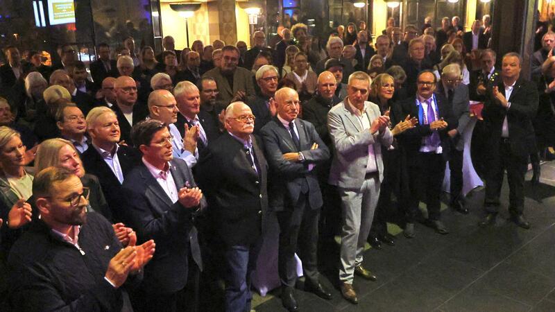 Große Gruppe von Menschen in formeller Kleidung, die in einem Innenraum stehen und applaudieren.